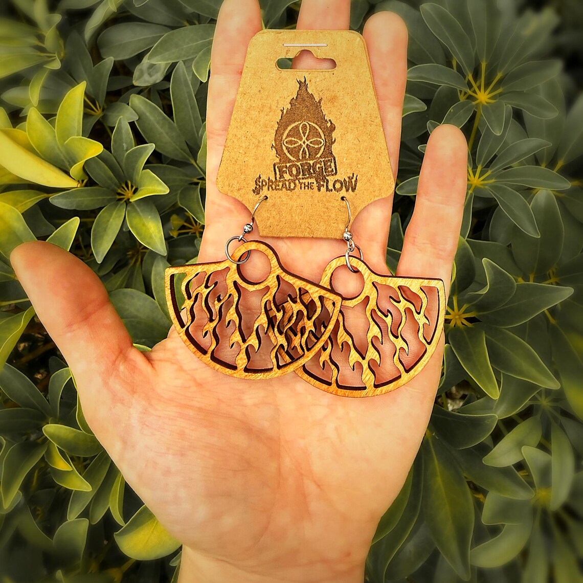 A pair of EMBER day fan earrings, miniature wooden flow fans with the same cut flame design, held in an open palm on a FORGE branded card