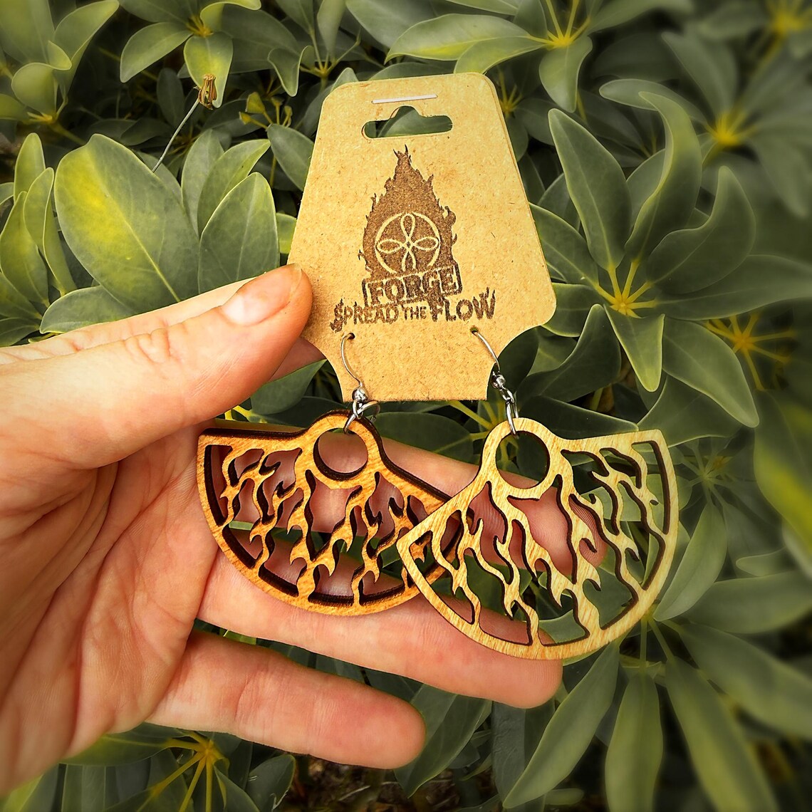 The earrings held between finger and thumb, showing the cut flame detail and the wood grain