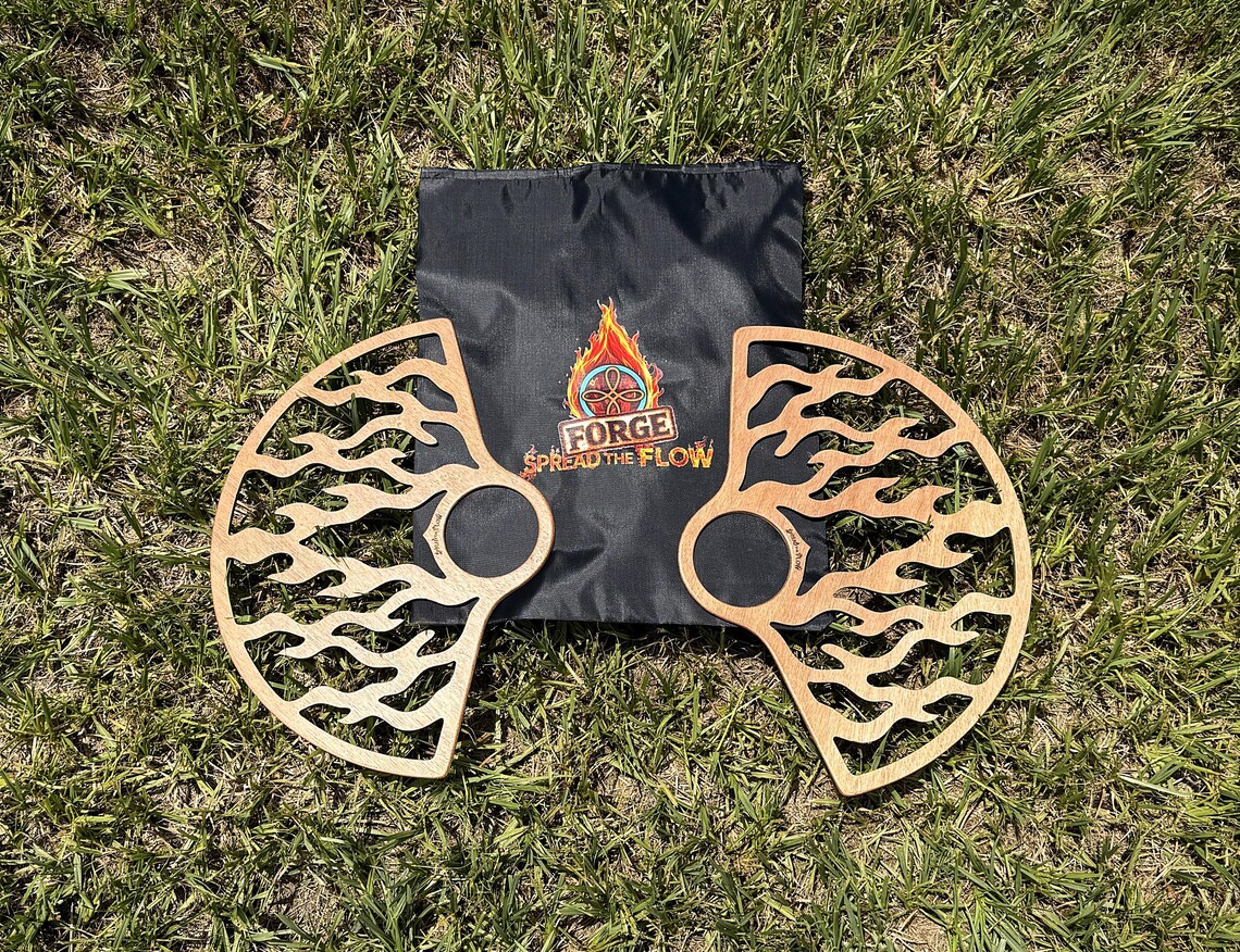 A pair of EMBER wooden flow fans with a cut flame design, laid on grass either side of a black FORGE drawstring carry bag